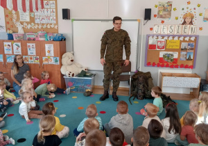 grupa dzieci siedzi na dywanie, umundurowany żołnierz opowiada o swojej profesji