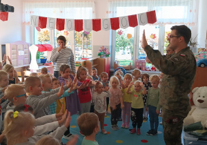 grupa dzieci stoi na dywanie, umundurowany żołnierz pokazuje gesty, które dzieci naśladuja