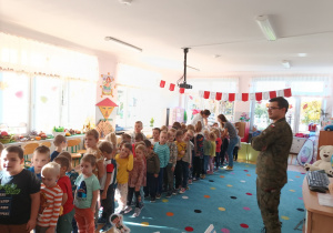 grupa dzieci stoi na dywanie, umundurowany żołnierz uczy dzieci "musztry"