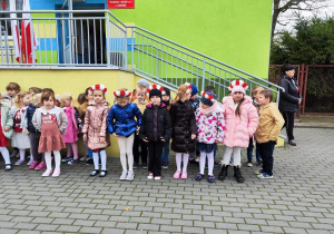 grupa dzieci przed budynkiem przedszkola, niektóre mają na głowach biało-czerwone wianki