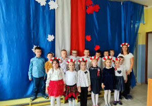 grupa dzieci i nauczycielka , wszyscy ubrani odświętnie pozują do zdjęcia na tle okolicznościowej biało-czerwonej dekoracji