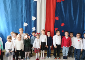grupa dzieci, ubranych na galowo, dziewczynki mają na głowach biało-czerwone wianki , w tle biało-czerwona dekoracja