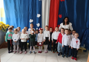 nauczycielka i grupa dzieci, wszyscy ubrani na galowo, dziewczynki mają na głowach biało-czerwone wianki , w tle biało-czerwona dekoracja