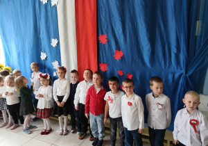 grupa dzieci, ubranych na galowo, dziewczynki mają na głowach biało-czerwone wianki , w tle biało-czerwona dekoracja