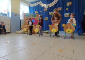 cztery dziewczynki biorą udział w zabawach sprawnościowych, przed nimi stoją krzesełka, na nich naklejone tekturowe głowy misiów, nauczycielka stoi za jedną z dziewczynek