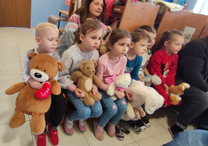 opiekunka i grupa dzieci siedzą w szatni na ławeczkach i kocach, w dłoniach trzymają misie