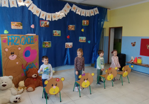 czworo dzieci bierze udział w zabawach sprawnościowych, przed nimi stoją krzesełka, na nich naklejone tekturowe głowy misiów,