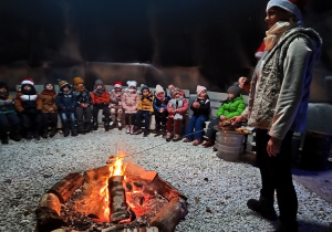 grupa dzieci siedzi w kręgu, w środku pali się ognisko, kobieta piecze w ognisku pianki