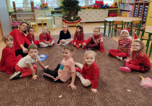 grupa dzieci i nauczycielka, siedzą w kole na dywanie mają na sobie mikołajkowe akcenty, w tle choinka