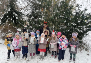 grupa dzieci i nauczycielka pozują do wspólnego zdjęcia, stoją przy "lasku" w przedszkolnym ogrodzie, jest śnieg