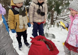 grupa dzieci w przedszkolnym ogrodzie z odnalezionym czerwonym workiem z prezentami, pochylają się nad nim