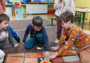 grupa dzieci w klasie, kucają na podłodze, tworzą wspólnie wielkoformatową kartę zabawy "kominek Mikołaja" - decydują gdzie umieścić "ognisko"