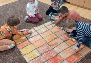 grupa dzieci w klasie, kucają na podłodze, tworzą wspólnie wielkoformatową kartę zabawy "kominek Mikołaja" - dwóch chłopców przecina po śladzie kartę