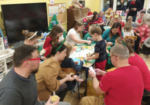 sala przedszkolna. grupa dzieci i opiekunów wspólnie przygotowuje kartki świąteczne. Niektórzy mają ubrania w świąteczne motywy lub mają na głowach świąteczne gadżety (czapki i rogi)