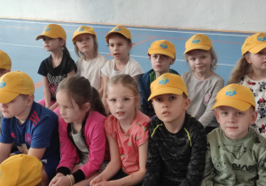 grupa dzieci w żółtych czapkach z daszkiem (z logo przedszkola) siedzi na ławkach na sali gimnastycznej w SP2