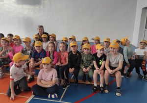 grupa dzieci w żółtych czapkach z daszkiem (z logo przedszkola) siedzi na ławkach na sali gimnastycznej w SP2