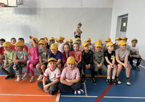 grupa dzieci w żółtych czapkach z logo przedszkola siedzi na ławkach w sali gimnastycznej SP2