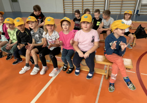 grupa dzieci w czapkach z logo przedszkola siedzi na ławce w sali gimnastycznej SP2