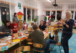 rodzice i dzieci siedzą przy stolikach , tworzą wspólnie kartki świateczne , obok stoi pani dyrektor