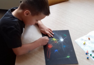 chłopiec siedzi przy stole i rozciera pastele na czarnej kartce
