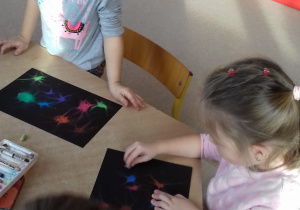 dziewczynki siedzą przy stole i rozcierają pastele na czarnej kartce