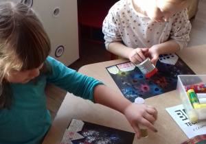 dwie dziewczynki posypują swoje gotowe pracy kolorowymi brokatami