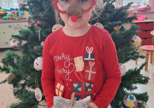 Dziewczynka pozuje do zdjęcia. Trzyma mikołajkowy prezent, a na twarzy ma okulary ze świątecznym akcentem. W tle znajduje się choinka.
