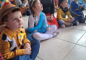 Dzieci w strojach karnawałowych siedzą w kole i słuchają wypowiedzi prowadzącego wydarzenie.