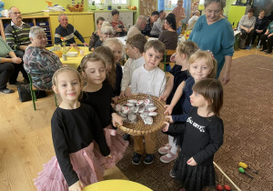 grupa dzieci z prezentami w koszyku, obok nauczycielka , zaproszeni na Dzień babci i Dziadka goście oraz dzieci na okolicznościowym poczęstunku w klasie