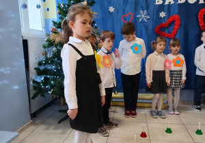 grupa dzieci ubranych odświętnie , za nimi okolicznościowa dekoracja, na podłodze przed dziećmi leżą dzwonki