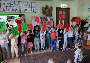 grupa dzieci unosi litery na zielonym i czerwonym tle, które układają się w napis: EKOLWAENTYNKI, obok stoją nauczycielki, w tle prezentacja związana z tematem