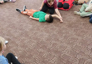 na dywanie leży chłopiec, drugi układa go w pozycji bezpiecznej, obok ratowniczka
