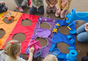 grupa dzieci obok nich kolorowa mata z otworami, dzieci segregują zabawki z sali wg kolorów maty