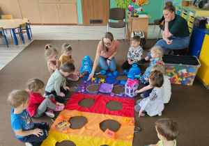 grupa dzieci obok nich kolorowa mata z otworami, dzieci segregują zabawki z sali wg kolorów maty, obok siedzą panie