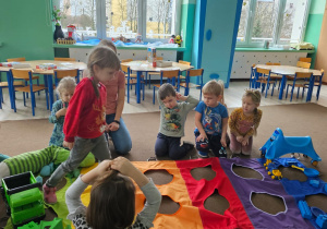 grupa dzieci obok nich kolorowa mata z otworami, dzieci segregują zabawki z sali wg kolorów maty