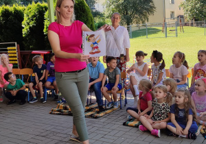 społeczność przedszkolna zebrana na tarasie , jedna z nauczycielek pokazuje ilustracje, dzieci odgadują z jakiej jest bajki