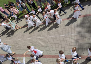 grupa dzieci tańczy poloneza , dziewczynki w białych sukienkach, przepasane czerwonymi wstążkami, we włosach czerwone kokardy. Chłopcy ubrani galowo. W oddali stoją rodzice