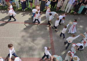 grupa dzieci tańczy poloneza , dziewczynki w białych sukienkach, przepasane czerwonymi wstążkami, we włosach czerwone kokardy. Chłopcy ubrani galowo. W oddali stoją rodzice