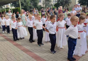 grupa dzieci tańczy poloneza , dziewczynki w białych sukienkach, przepasane czerwonymi wstążkami, we włosach czerwone kokardy. Chłopcy ubrani galowo. W oddali stoją rodzice