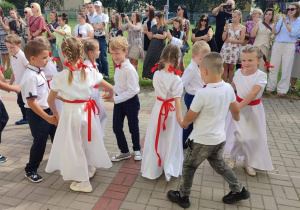 grupa dzieci tańczy poloneza , dziewczynki w białych sukienkach, przepasane czerwonymi wstążkami, we włosach czerwone kokardy. Chłopcy ubrani galowo. W oddali stoją rodzice