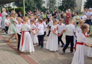 grupa dzieci tańczy poloneza , dziewczynki w białych sukienkach, przepasane czerwonymi wstążkami, we włosach czerwone kokardy. Chłopcy ubrani galowo. W oddali stoją rodzice