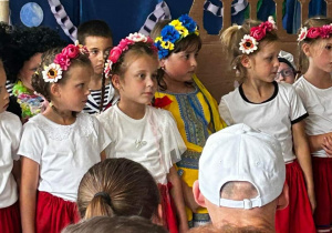 grupa dzieci na scenie, dziewczynki ubrane na biało-czerwono, na głowach mają kolorowe wianki , jedna z dziewczynek ubrana w strój ukraiński