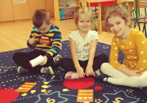 Dzieci siedzą na dywanie i grają w grę "kurnik", dwie dziewczynki patrzą w obiektyw i uśmiechają się