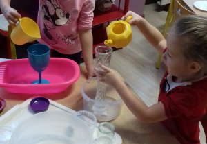dwie dziewczynki przy stole przelewają wodę z różnych naczyń
