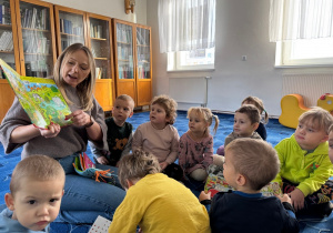 dzieci siedzą na dywanie w bibliotece, przy nich pani bibliotekarka, która pokazuje dzieciom książkę