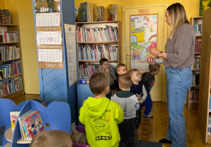 dzieci w bibliotece, pani bibliotekarka pokazuje im ksiażeczki umieszczone na regałach