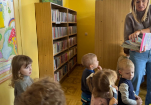 dzieci w bibliotece, pani bibliotekarka pokazuje im ksiażeczki i opowiada o zasadach zachowania się w bibliotece