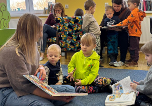 dzieci siedzą na dywanie w bibliotece, przy nich pani bibliotekarka, przed którą leżą książki , na drugim planie inne dzieci i panie oglądają ksiażeczki