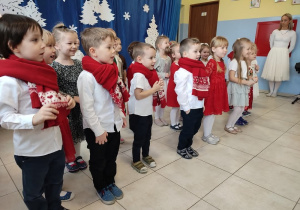 dzieci prezentują świąteczny występ, są odświętnie ubrane , niektóre mają na szyjach czerwone szaliki