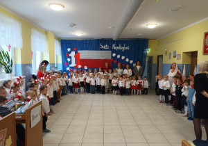 dzieci i nauczycielki w biaLo-czerwonych ubraniach, ustawieni w kole w szatni, przygotowują się do śpiewania Mazurka Dąbrowskiego, w tle patriotyczna dekoracja